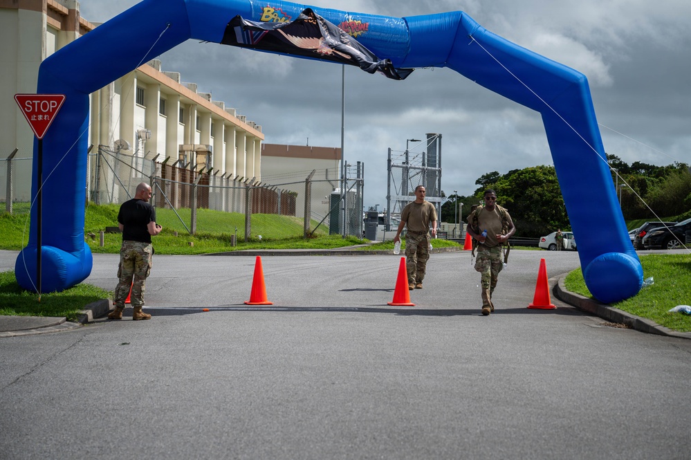 Military personnel are approaching a finish line marked by a large blue inflatable arch on a paved road at what appears to be a military installation. Two participants in camouflage uniforms and carrying gear are walking through the arch while a third person in black and camouflage stands to the side, likely as an official or monitor. The setting includes orange traffic cones, a bilingual stop sign in English and Japanese, and military base infrastructure in the background.