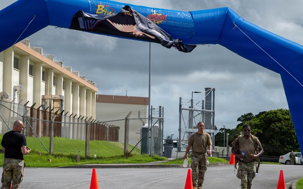 Military personnel are approaching a finish line marked by a large blue inflatable arch on a paved road at what appears to be a military installation. Two participants in camouflage uniforms and carrying gear are walking through the arch while a third person in black and camouflage stands to the side, likely as an official or monitor. The setting includes orange traffic cones, a bilingual stop sign in English and Japanese, and military base infrastructure in the background.