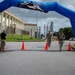 Military personnel are approaching a finish line marked by a large blue inflatable arch on a paved road at what appears to be a military installation. Two participants in camouflage uniforms and carrying gear are walking through the arch while a third person in black and camouflage stands to the side, likely as an official or monitor. The setting includes orange traffic cones, a bilingual stop sign in English and Japanese, and military base infrastructure in the background.