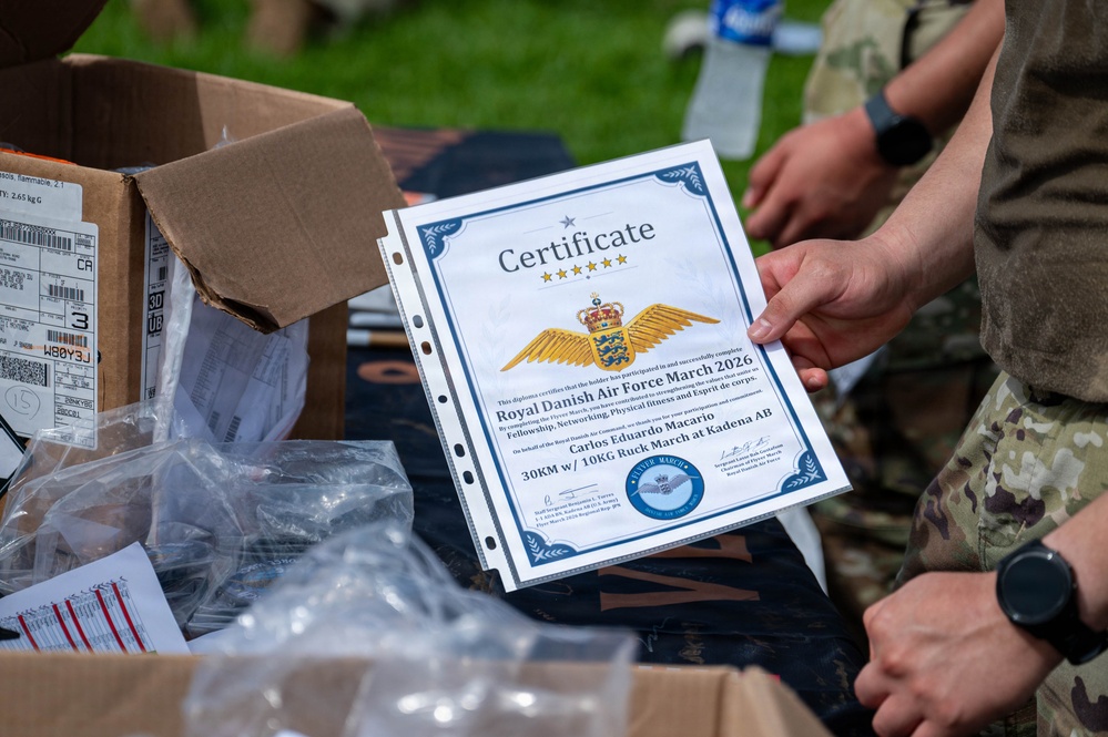 A military certificate from the Royal Danish Air Force March 2026 is being held up for display. The certificate is awarded to Carlos Eduardo Macario for completing a 30KM march with a 10KG ruck at Kadena AB. The setting appears to be an outdoor distribution or awards table with boxes and supplies visible.