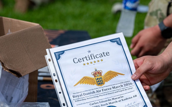 A military certificate from the Royal Danish Air Force March 2026 is being held up for display. The certificate is awarded to Carlos Eduardo Macario for completing a 30KM march with a 10KG ruck at Kadena AB. The setting appears to be an outdoor distribution or awards table with boxes and supplies visible.