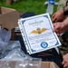 A military certificate from the Royal Danish Air Force March 2026 is being held up for display. The certificate is awarded to Carlos Eduardo Macario for completing a 30KM march with a 10KG ruck at Kadena AB. The setting appears to be an outdoor distribution or awards table with boxes and supplies visible.