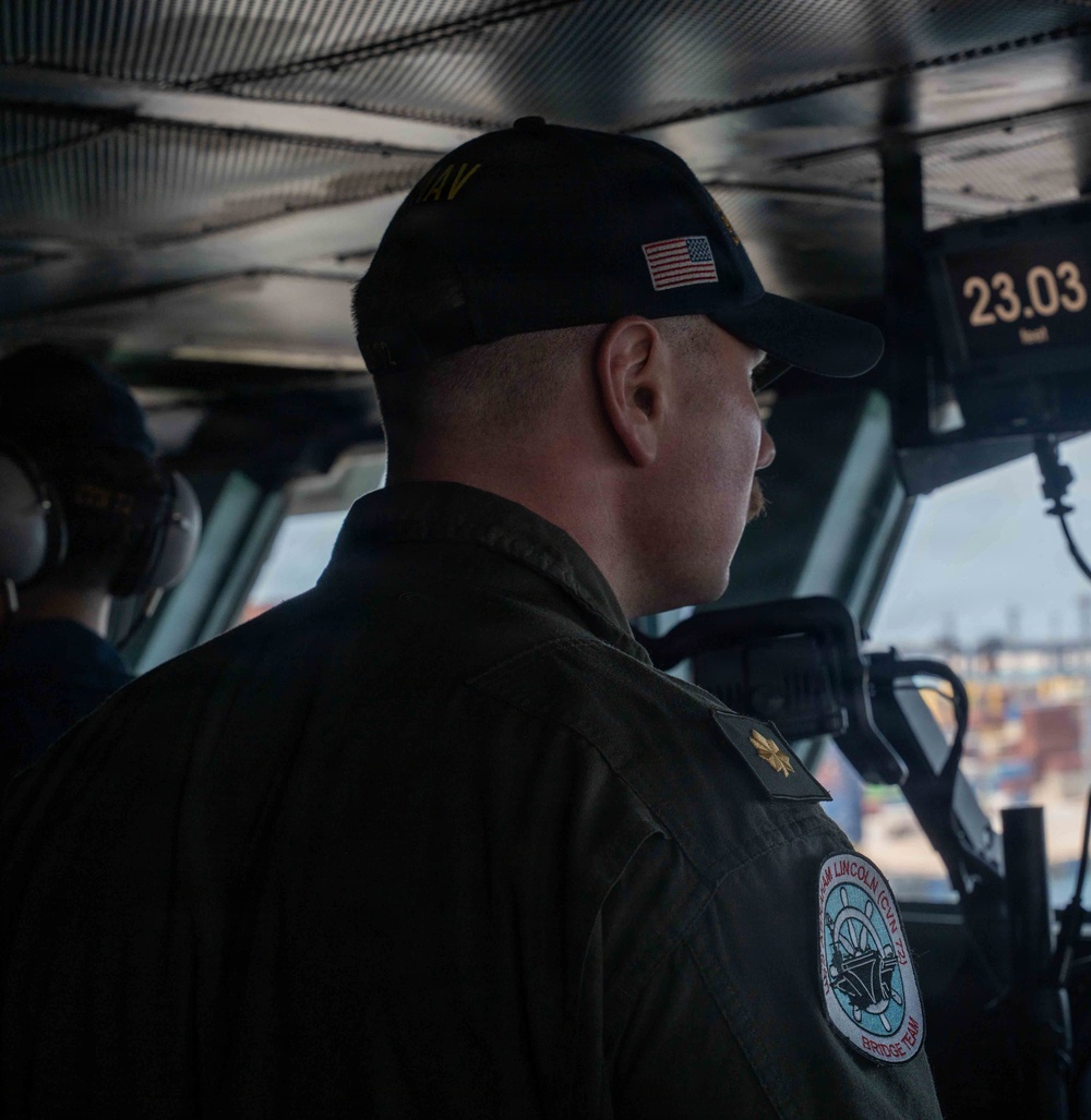 A U.S. Navy officer is photographed from behind on the bridge of USS Abraham Lincoln (CVN-72), wearing a dark ball cap with an American flag patch and a flight deck jacket with a 'USS Abraham Lincoln (CVN-72) Bridge Team' patch. A gold oak leaf insignia is visible on the shoulder, indicating the rank of Lieutenant Commander or Major. A digital display reading '23.03' is visible in the upper right, and port infrastructure is visible through the bridge windows.