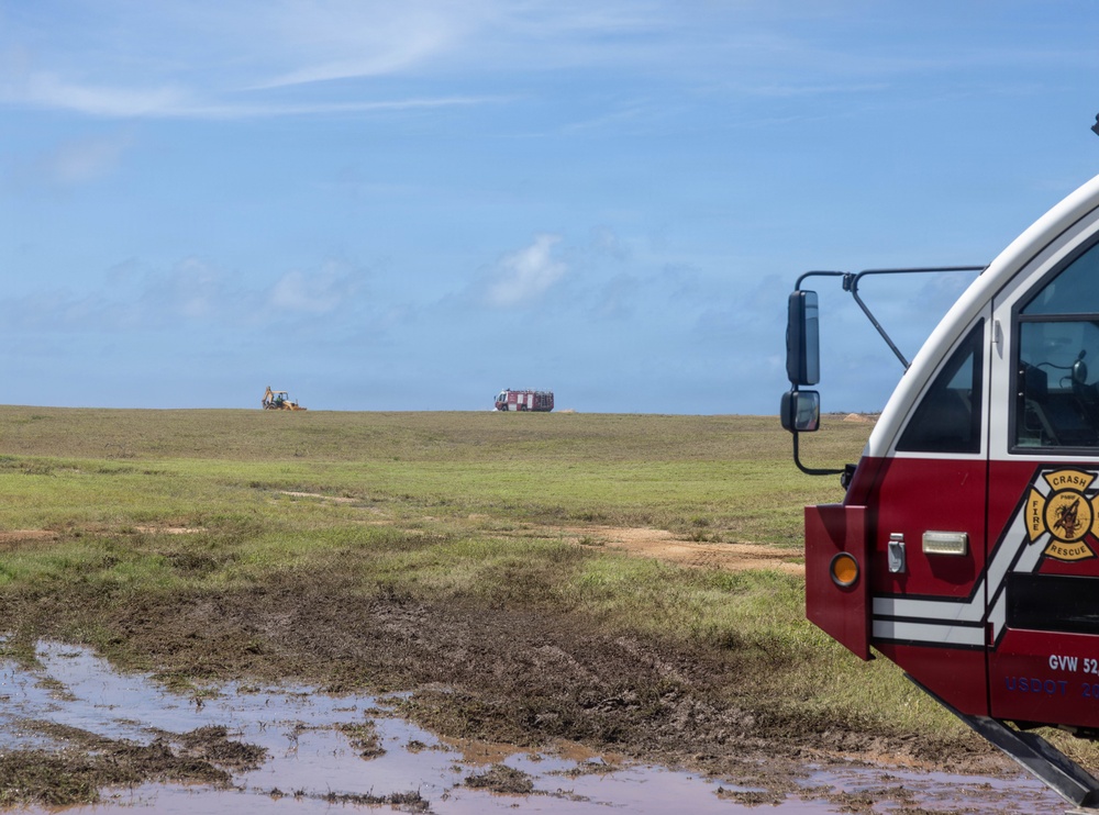 A Crash Fire Rescue (CFR) vehicle is prominently visible in the foreground right of the image, positioned on muddy terrain near an open grassy field. In the distant background, a second fire/rescue truck and what appears to be a yellow construction excavator are visible on the horizon. The scene suggests emergency standby operations, possibly at an airfield or large open facility.