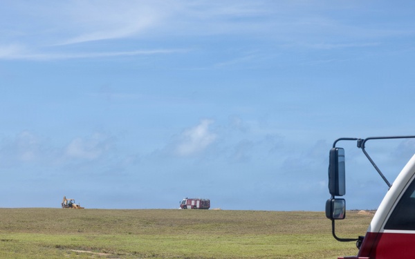 A Crash Fire Rescue (CFR) vehicle is prominently visible in the foreground right of the image, positioned on muddy terrain near an open grassy field. In the distant background, a second fire/rescue truck and what appears to be a yellow construction excavator are visible on the horizon. The scene suggests emergency standby operations, possibly at an airfield or large open facility.