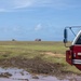 A Crash Fire Rescue (CFR) vehicle is prominently visible in the foreground right of the image, positioned on muddy terrain near an open grassy field. In the distant background, a second fire/rescue truck and what appears to be a yellow construction excavator are visible on the horizon. The scene suggests emergency standby operations, possibly at an airfield or large open facility.