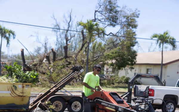 A worker in a bright green shirt operates a Ditch Witch skid steer loader during post-storm debris cleanup operations. The foreground shows an uprooted tree with exposed root ball and scattered branches, while the background reveals heavily damaged and stripped trees consistent with major storm damage. A yellow dumpster and white pickup truck are also visible in the staging area.