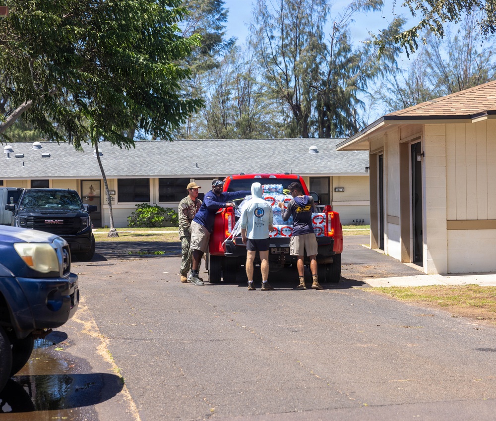 Four individuals are unloading cases of bottled water from the bed of a red pickup truck in a residential or institutional parking area. One person appears to be wearing military-style camouflage clothing, suggesting possible disaster relief or community aid activity. The setting includes low single-story buildings and tall trees under a clear blue sky.