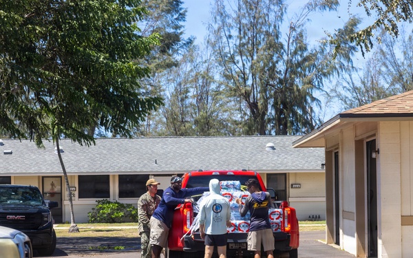 Four individuals are unloading cases of bottled water from the bed of a red pickup truck in a residential or institutional parking area. One person appears to be wearing military-style camouflage clothing, suggesting possible disaster relief or community aid activity. The setting includes low single-story buildings and tall trees under a clear blue sky.
