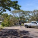 A US Navy pickup truck is parked near a fallen tree that is blocking or partially obstructing a road or parking area. A worker appears to be using a cable or rope, likely attached to the truck, to assist in removing the downed tree. The scene suggests storm damage cleanup operations at what appears to be a military installation.