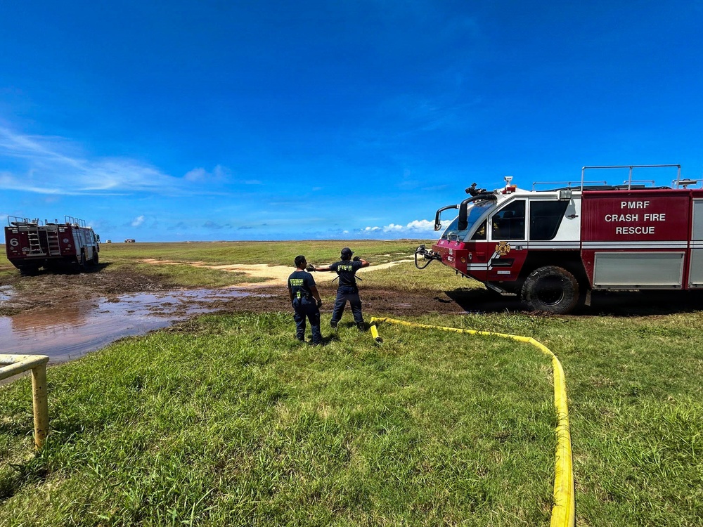Two firefighters from PMRF (Pacific Missile Range Facility) Crash Fire Rescue are operating on an open grassy field, managing a yellow fire hose connected to a red and white crash fire rescue truck. A second fire truck, numbered '6', is visible in the background near a flooded or muddy area. The scene appears to be a training exercise or emergency response operation under clear blue skies.