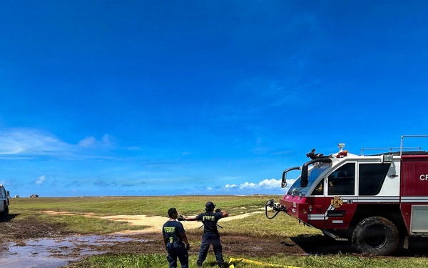 Two firefighters from PMRF (Pacific Missile Range Facility) Crash Fire Rescue are operating on an open grassy field, managing a yellow fire hose connected to a red and white crash fire rescue truck. A second fire truck, numbered '6', is visible in the background near a flooded or muddy area. The scene appears to be a training exercise or emergency response operation under clear blue skies.