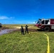 Two firefighters from PMRF (Pacific Missile Range Facility) Crash Fire Rescue are operating on an open grassy field, managing a yellow fire hose connected to a red and white crash fire rescue truck. A second fire truck, numbered '6', is visible in the background near a flooded or muddy area. The scene appears to be a training exercise or emergency response operation under clear blue skies.