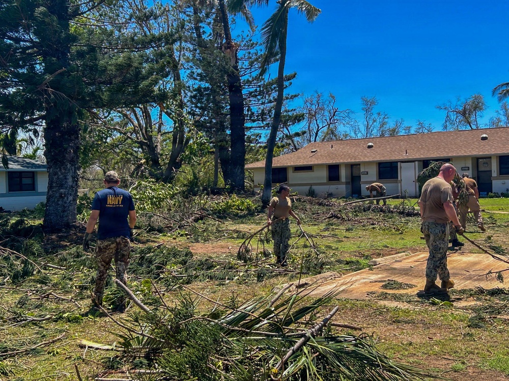 Military personnel, including at least one individual wearing a U.S. Navy t-shirt, are conducting storm debris cleanup operations around residential or barracks-style buildings. The area shows significant storm damage with numerous downed trees, broken branches, and scattered palm fronds covering the ground. The clear blue sky and tropical vegetation suggest a post-hurricane or typhoon recovery effort at a military installation.