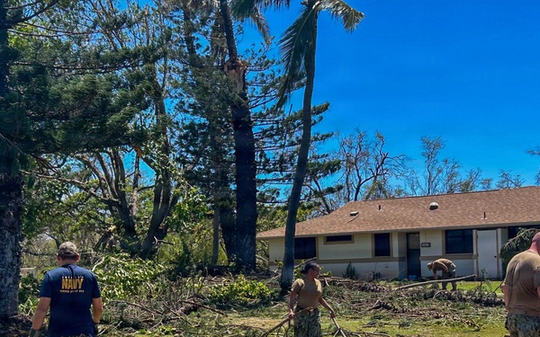 Military personnel, including at least one individual wearing a U.S. Navy t-shirt, are conducting storm debris cleanup operations around residential or barracks-style buildings. The area shows significant storm damage with numerous downed trees, broken branches, and scattered palm fronds covering the ground. The clear blue sky and tropical vegetation suggest a post-hurricane or typhoon recovery effort at a military installation.