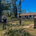 Military personnel, including at least one individual wearing a U.S. Navy t-shirt, are conducting storm debris cleanup operations around residential or barracks-style buildings. The area shows significant storm damage with numerous downed trees, broken branches, and scattered palm fronds covering the ground. The clear blue sky and tropical vegetation suggest a post-hurricane or typhoon recovery effort at a military installation.