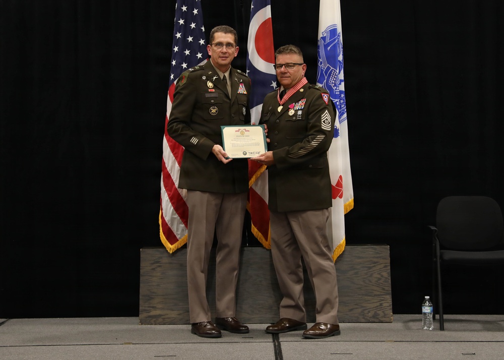 Two U.S. Army soldiers in dress green uniforms stand on a stage holding a certificate or commendation document together. The ceremony appears to be a formal military awards or retirement event, with the soldier on the right displaying multiple medals and a distinctive red-and-gold neck decoration. Three flags are visible in the background, including the U.S. flag, the Ohio state flag, and what appears to be a military unit or command flag.
