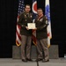 Two U.S. Army soldiers in dress green uniforms stand on a stage holding a certificate or commendation document together. The ceremony appears to be a formal military awards or retirement event, with the soldier on the right displaying multiple medals and a distinctive red-and-gold neck decoration. Three flags are visible in the background, including the U.S. flag, the Ohio state flag, and what appears to be a military unit or command flag.