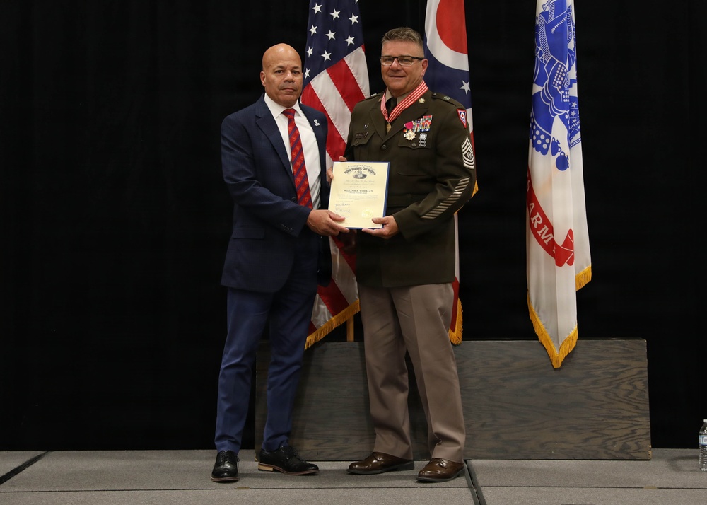 Two men are posing together on a stage holding a framed certificate or award document. One man is wearing a U.S. Army dress uniform with multiple medals and ribbons, along with a red lanyard, while the other is dressed in a civilian navy blue suit with a red tie. The ceremony appears to be a military recognition or retirement event, with American, Ohio state, and U.S. Army flags visible in the background.