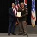 Two men are posing together on a stage holding a framed certificate or award document. One man is wearing a U.S. Army dress uniform with multiple medals and ribbons, along with a red lanyard, while the other is dressed in a civilian navy blue suit with a red tie. The ceremony appears to be a military recognition or retirement event, with American, Ohio state, and U.S. Army flags visible in the background.