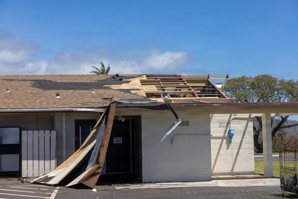 A single-story commercial or institutional building has sustained significant wind damage, with large sections of roofing material and sheathing torn away, exposing the underlying wooden roof framing. Debris including roofing panels and underlayment has peeled back and collapsed against the front facade near the entrance door. The damage is consistent with hurricane or severe windstorm impact.