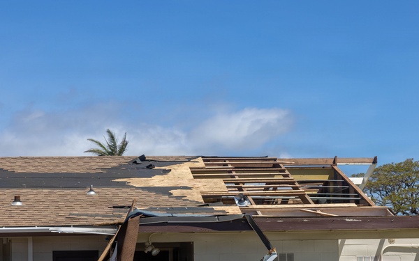 A single-story commercial or institutional building has sustained significant wind damage, with large sections of roofing material and sheathing torn away, exposing the underlying wooden roof framing. Debris including roofing panels and underlayment has peeled back and collapsed against the front facade near the entrance door. The damage is consistent with hurricane or severe windstorm impact.