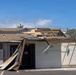 A single-story commercial or institutional building has sustained significant wind damage, with large sections of roofing material and sheathing torn away, exposing the underlying wooden roof framing. Debris including roofing panels and underlayment has peeled back and collapsed against the front facade near the entrance door. The damage is consistent with hurricane or severe windstorm impact.