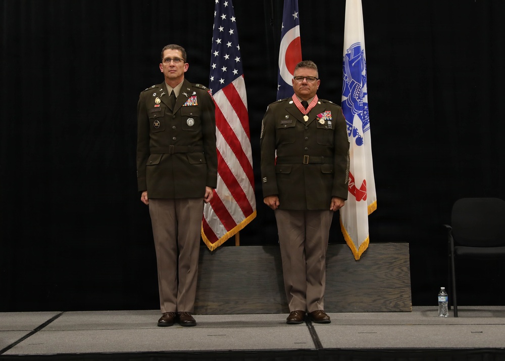 Two military personnel in U.S. Army dress green uniforms stand at attention on a stage during what appears to be a formal military ceremony. The individual on the right is wearing a large medal with a red ribbon around his neck, suggesting he is being honored or has recently received an award. Three flags are displayed behind them: the U.S. flag, the Ohio state flag, and what appears to be a U.S. Coast Guard or similar organizational flag.