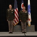 Two military personnel in U.S. Army dress green uniforms stand at attention on a stage during what appears to be a formal military ceremony. The individual on the right is wearing a large medal with a red ribbon around his neck, suggesting he is being honored or has recently received an award. Three flags are displayed behind them: the U.S. flag, the Ohio state flag, and what appears to be a U.S. Coast Guard or similar organizational flag.