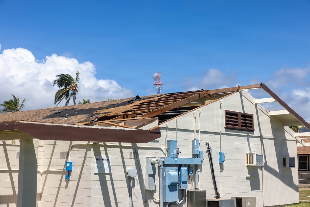 A commercial or institutional building has sustained significant roof damage, with large sections of roofing material and sheathing torn away, exposing the underlying wooden rafters and decking. The damage appears consistent with high-wind event such as a hurricane or severe storm, supported by the visibly wind-stressed palm tree in the background. The building's exterior walls remain intact, and electrical utility equipment is mounted on the exterior facade.