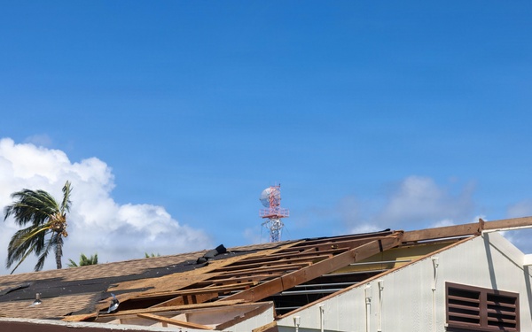 A commercial or institutional building has sustained significant roof damage, with large sections of roofing material and sheathing torn away, exposing the underlying wooden rafters and decking. The damage appears consistent with high-wind event such as a hurricane or severe storm, supported by the visibly wind-stressed palm tree in the background. The building's exterior walls remain intact, and electrical utility equipment is mounted on the exterior facade.