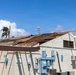 A commercial or institutional building has sustained significant roof damage, with large sections of roofing material and sheathing torn away, exposing the underlying wooden rafters and decking. The damage appears consistent with high-wind event such as a hurricane or severe storm, supported by the visibly wind-stressed palm tree in the background. The building's exterior walls remain intact, and electrical utility equipment is mounted on the exterior facade.