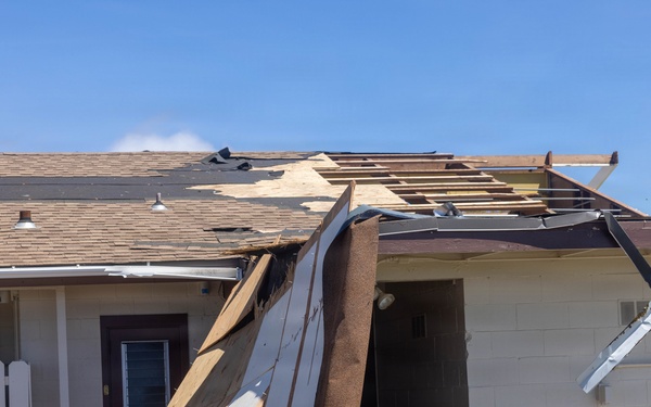 A commercial or institutional building has sustained significant roof damage, with large sections of roofing material, sheathing, and metal flashing peeled away and hanging down toward the ground. The exposed wooden roof framing (rafters and decking) is clearly visible where the covering has been torn off. The damage pattern is consistent with high-wind event such as a hurricane or severe storm.