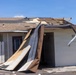 A commercial or institutional building has sustained significant roof damage, with large sections of roofing material, sheathing, and metal flashing peeled away and hanging down toward the ground. The exposed wooden roof framing (rafters and decking) is clearly visible where the covering has been torn off. The damage pattern is consistent with high-wind event such as a hurricane or severe storm.