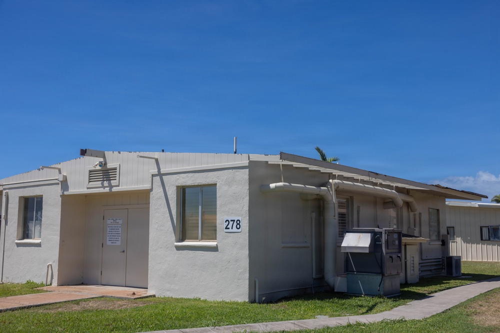 A single-story, white stucco building numbered '278' photographed under a clear blue sky. The structure appears to be a utilitarian or institutional building, possibly located on a military base or government facility, featuring minimal architectural ornamentation and visible HVAC equipment. The surrounding grounds are well-maintained with trimmed grass and a concrete walkway.