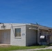 A single-story, white stucco building numbered '278' photographed under a clear blue sky. The structure appears to be a utilitarian or institutional building, possibly located on a military base or government facility, featuring minimal architectural ornamentation and visible HVAC equipment. The surrounding grounds are well-maintained with trimmed grass and a concrete walkway.