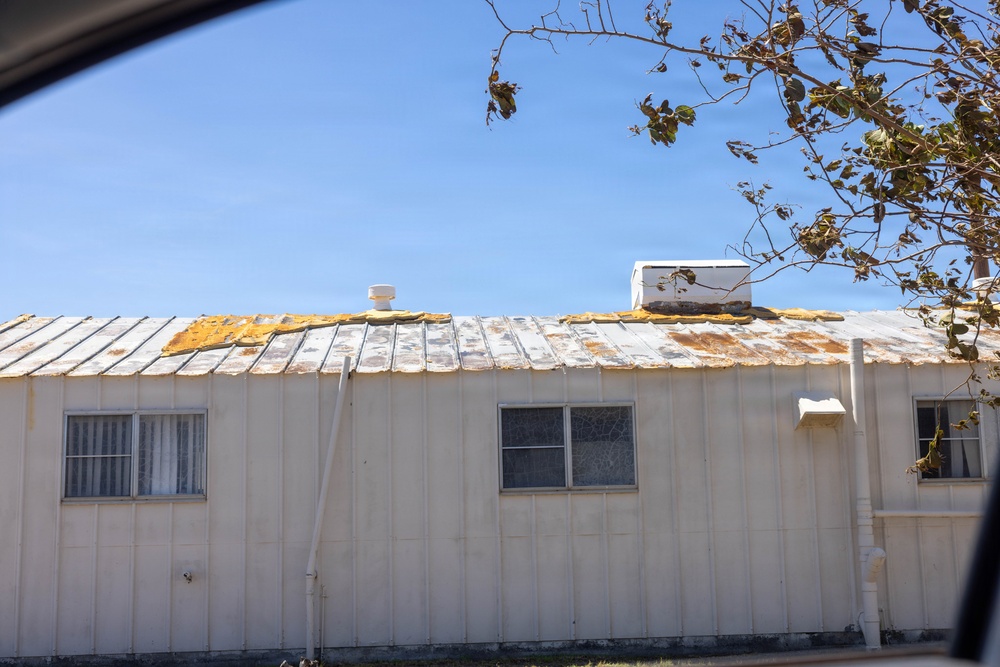 The image shows a deteriorating single-story building, likely a mobile home or modular structure, photographed from inside a vehicle. The metal roof shows significant damage with peeling paint, rust stains, and exposed insulation or underlayment material in yellow patches. The building appears aged and poorly maintained, with weathered white siding and windows showing signs of neglect.