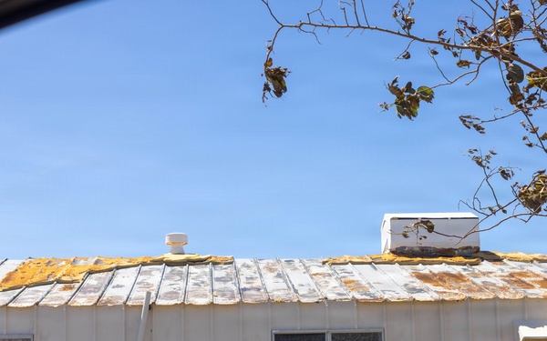 The image shows a deteriorating single-story building, likely a mobile home or modular structure, photographed from inside a vehicle. The metal roof shows significant damage with peeling paint, rust stains, and exposed insulation or underlayment material in yellow patches. The building appears aged and poorly maintained, with weathered white siding and windows showing signs of neglect.