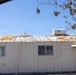 The image shows a deteriorating single-story building, likely a mobile home or modular structure, photographed from inside a vehicle. The metal roof shows significant damage with peeling paint, rust stains, and exposed insulation or underlayment material in yellow patches. The building appears aged and poorly maintained, with weathered white siding and windows showing signs of neglect.
