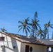The image shows a building with significant storm or wind damage to its roof, with shingles torn away and roofing material visibly peeled back. The structure appears to be located in a tropical or subtropical environment, evidenced by the palm trees and Norfolk Island pines visible in the background. The damage suggests the aftermath of a severe weather event such as a hurricane or strong windstorm.