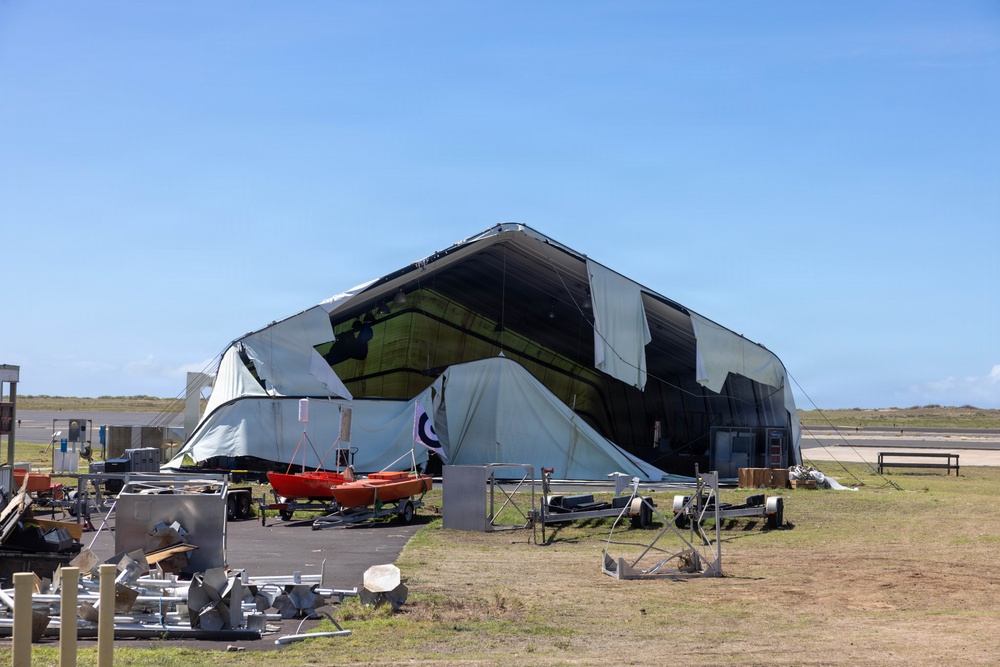 A large fabric or tensile structure, likely a temporary hangar or event tent, has suffered significant structural damage — its covering is torn, collapsed, and billowing outward. The surrounding area shows scattered metal debris, equipment, and small watercraft, suggesting storm or wind damage. The scene appears to be located near an airfield or coastal facility based on the flat terrain and runway visible in the background.