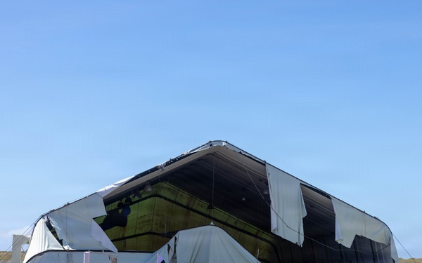 A large fabric or tensile structure, likely a temporary hangar or event tent, has suffered significant structural damage — its covering is torn, collapsed, and billowing outward. The surrounding area shows scattered metal debris, equipment, and small watercraft, suggesting storm or wind damage. The scene appears to be located near an airfield or coastal facility based on the flat terrain and runway visible in the background.