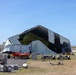 A large fabric or tensile structure, likely a temporary hangar or event tent, has suffered significant structural damage — its covering is torn, collapsed, and billowing outward. The surrounding area shows scattered metal debris, equipment, and small watercraft, suggesting storm or wind damage. The scene appears to be located near an airfield or coastal facility based on the flat terrain and runway visible in the background.
