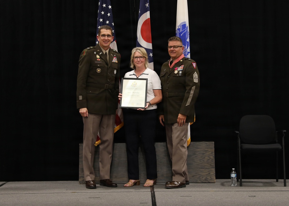 Three individuals pose for a formal photograph on a stage during what appears to be a military ceremony or award presentation. A civilian woman in the center holds a framed certificate or commendation, flanked by two U.S. Army soldiers in dress uniforms. The backdrop includes the American flag, the Ohio state flag, and what appears to be a Coast Guard or similar service flag.