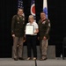 Three individuals pose for a formal photograph on a stage during what appears to be a military ceremony or award presentation. A civilian woman in the center holds a framed certificate or commendation, flanked by two U.S. Army soldiers in dress uniforms. The backdrop includes the American flag, the Ohio state flag, and what appears to be a Coast Guard or similar service flag.