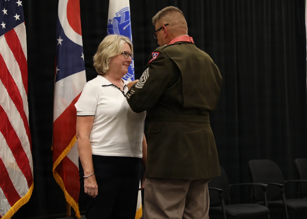 A military ceremony is taking place in which a uniformed soldier is pinning a medal or insignia onto a civilian woman in a white polo shirt. The setting includes the U.S. flag and the Ohio state flag in the background, suggesting this is an Ohio National Guard or Ohio-based military event. The woman is smiling, indicating a positive recognition or award ceremony.