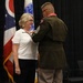A military ceremony is taking place in which a uniformed soldier is pinning a medal or insignia onto a civilian woman in a white polo shirt. The setting includes the U.S. flag and the Ohio state flag in the background, suggesting this is an Ohio National Guard or Ohio-based military event. The woman is smiling, indicating a positive recognition or award ceremony.