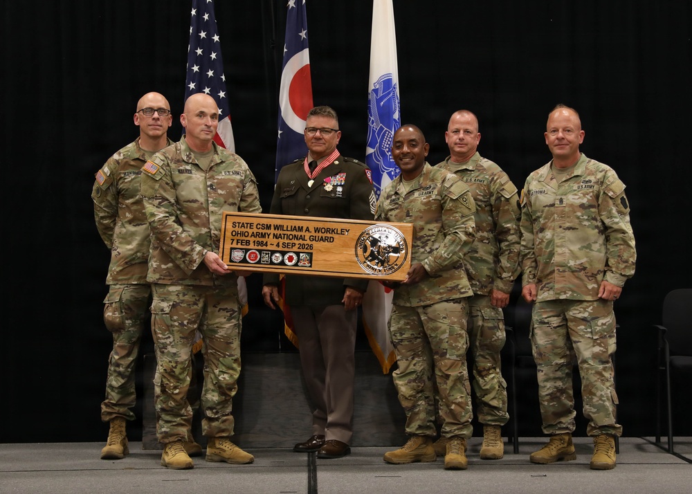 A military ceremony honoring State Command Sergeant Major William A. Workley of the Ohio Army National Guard, with six military personnel posing together holding a commemorative wooden plaque. The plaque marks his service dates from 7 February 1984 to 4 September 2026. Five individuals are in U.S. Army camouflage uniforms while one is in a Marine Corps dress uniform adorned with medals.