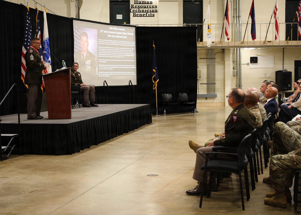 A military ceremony or presentation is taking place in what appears to be a National Guard armory or similar facility. A soldier is speaking at a podium on a raised stage, with a projection screen displaying information about 'CSM William Wordley, 10th State Command Sergeant Major (Retired), Ohio Army National Guard.' An audience of military personnel and civilians is seated in chairs facing the stage.