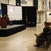 A military ceremony or presentation is taking place in what appears to be a National Guard armory or similar facility. A soldier is speaking at a podium on a raised stage, with a projection screen displaying information about 'CSM William Wordley, 10th State Command Sergeant Major (Retired), Ohio Army National Guard.' An audience of military personnel and civilians is seated in chairs facing the stage.